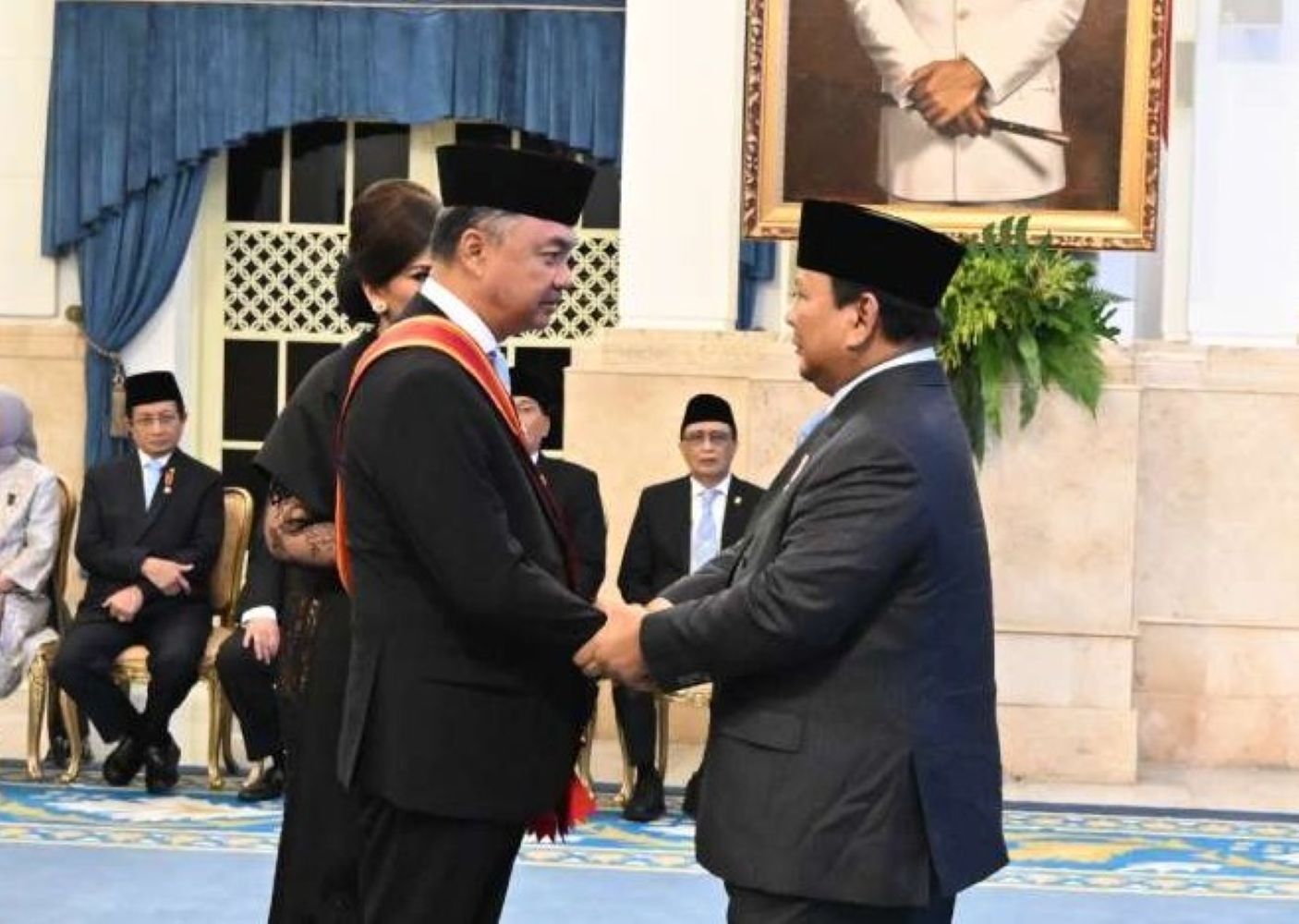 Two men in suits wearing traditional black caps shake hands during a formal ceremony with seated officials in the background and a portrait on the wall