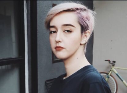Portrait of a person with short pastel pink and blonde hair standing near a doorway with a bicycle in the background