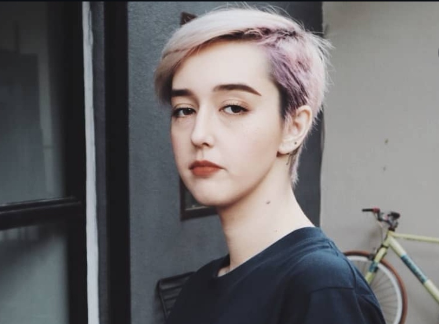 Portrait of a person with short pastel pink and blonde hair standing near a doorway with a bicycle in the background