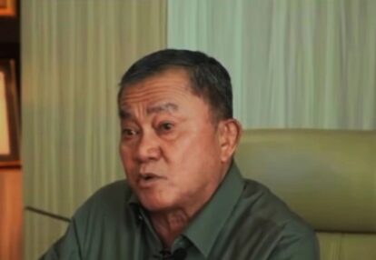 Man in a dark green shirt seated indoors with pale green curtains in the background