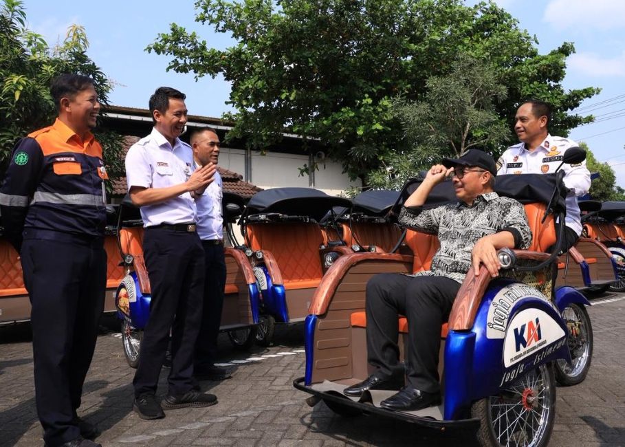 Man in a cap sits in a blue and orange pedicab while four uniformed men chat beside a row of orange rickshaws in a sunny parking lot