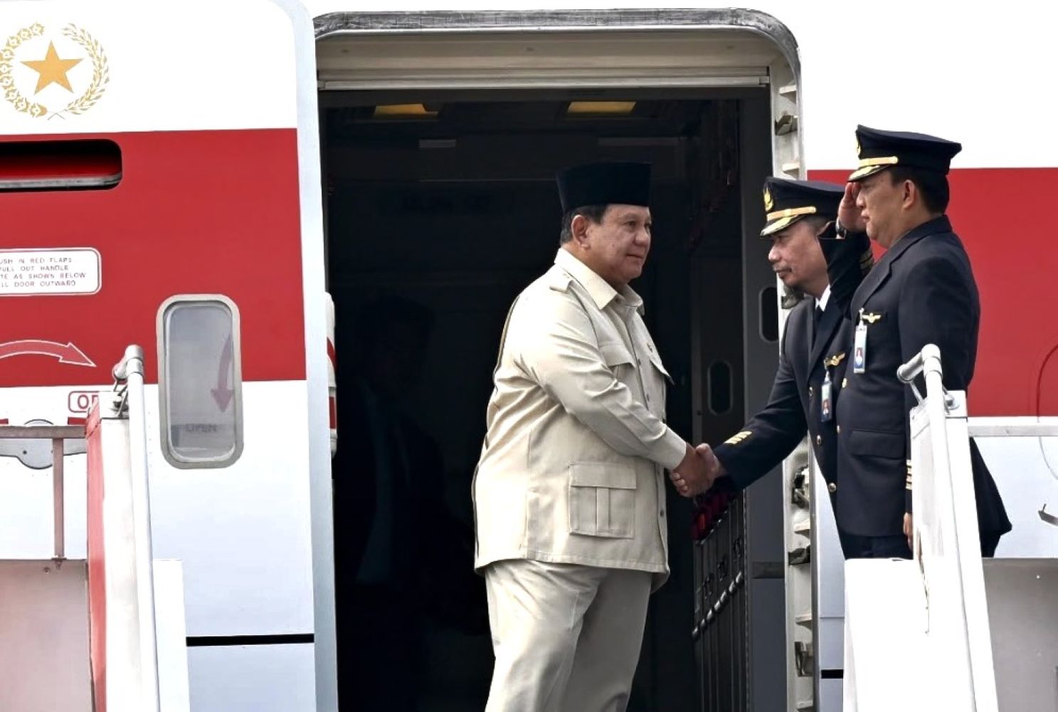 Two men shake hands at the open door of a red and white airplane one in a beige suit and black cap the other in a navy pilot uniform with gold badges