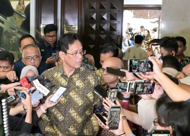 Man in a batik shirt speaks to reporters surrounded by microphones and smartphones in a crowded lobby corridor in front of a wooden door.