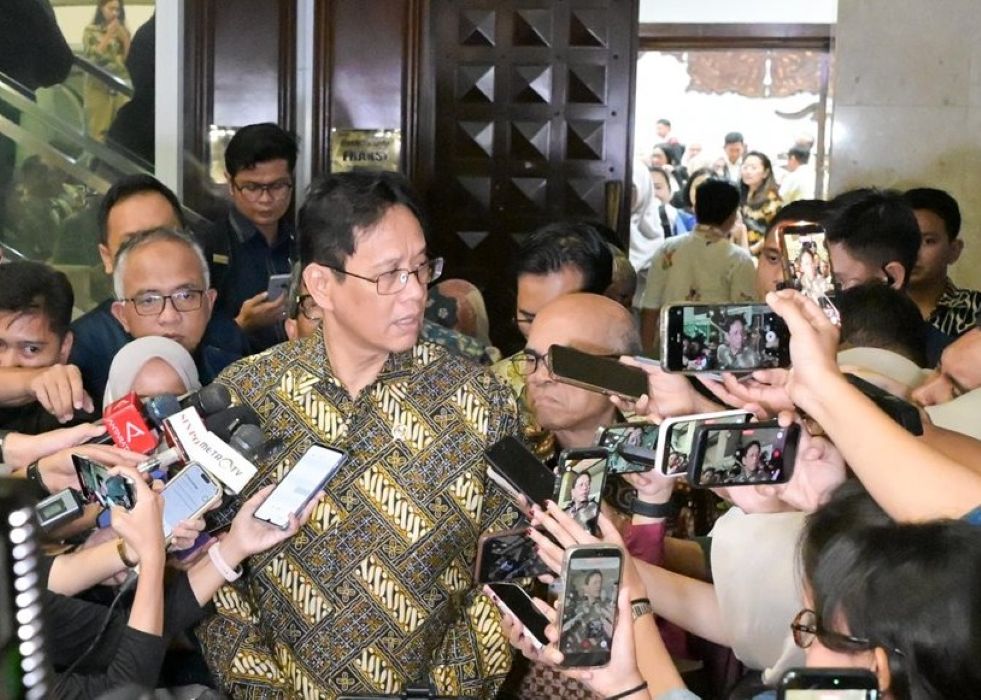 Man in a batik shirt speaks to reporters surrounded by microphones and smartphones in a crowded lobby corridor in front of a wooden door