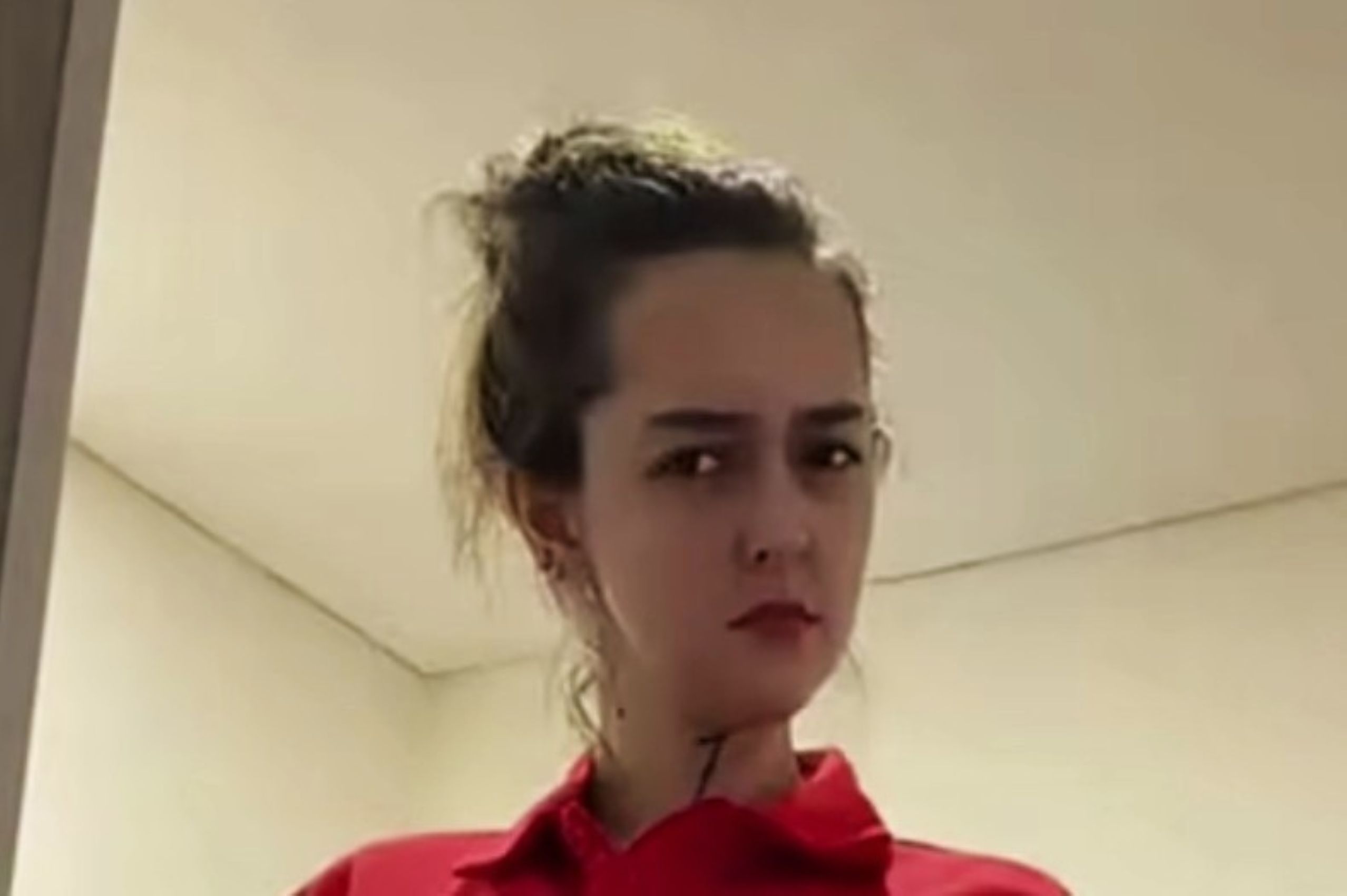 Close up of a person with dark hair tied back wearing a red shirt looking to the side in an indoor setting