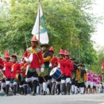 Parade of people in red uniforms marching with drums and a flag down a tree lined street with spectators nearby