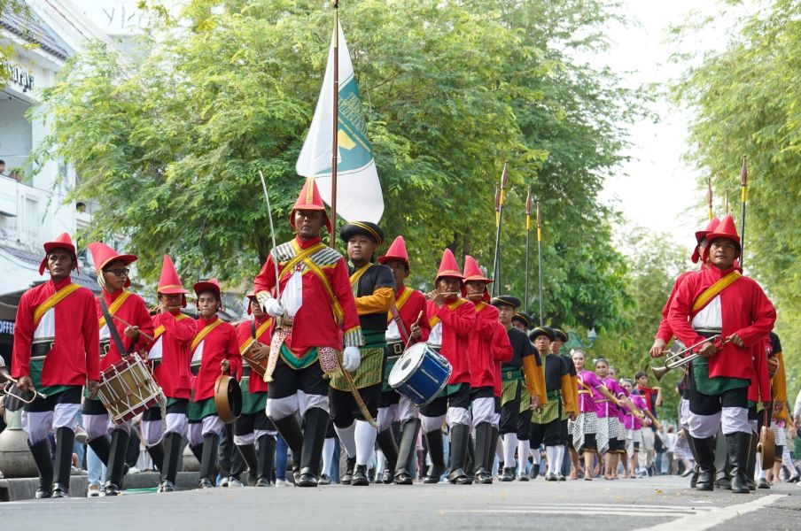 7fkirab catrawarta Parade of people in red uniforms marching with drums and a flag down a tree lined street with spectators nearby