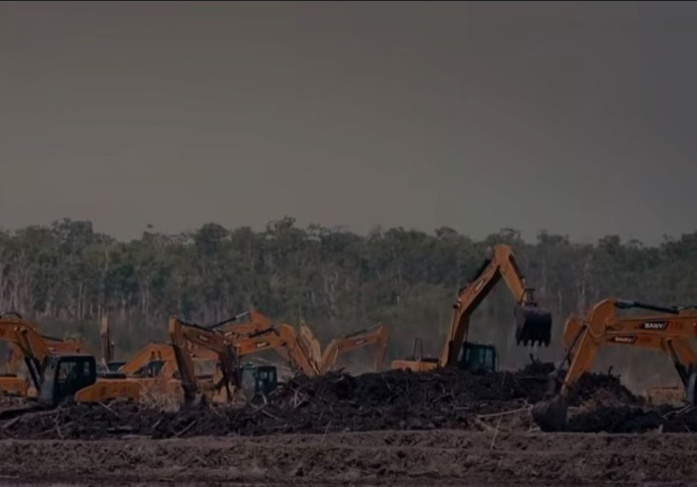Several orange excavators work on a dirt site with a large pile of debris in the foreground trees in the background