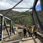 Group of construction workers in hard hats and yellow vests stand on a partially completed steel bridge with a forested hillside in the background