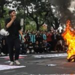 A young man in black speaks into a microphone as a pile of tires burns on a street protest with a large crowd gathered behind him