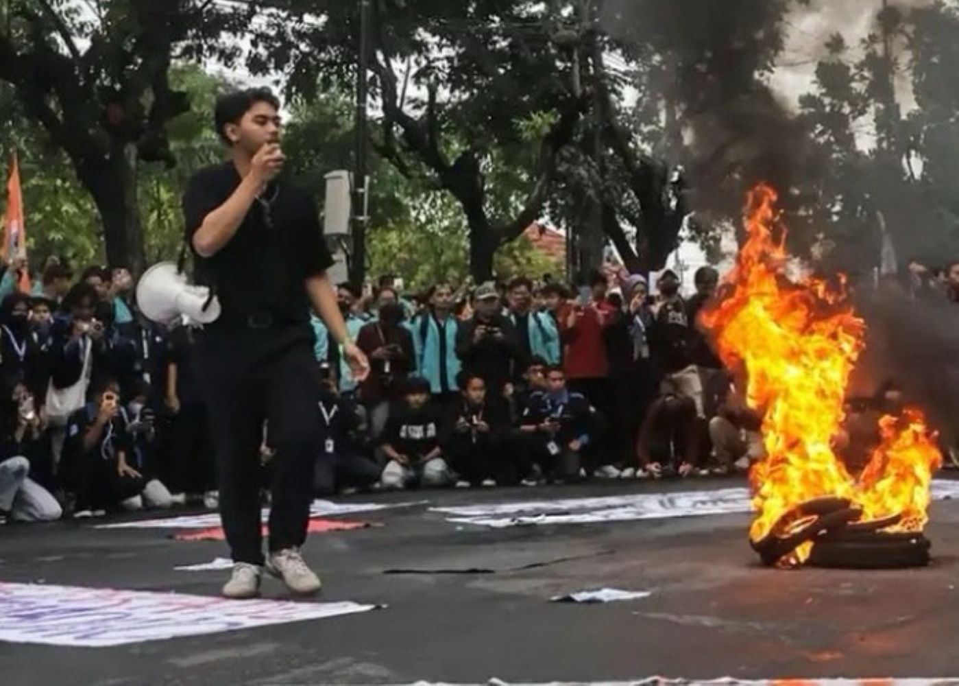 A young man in black speaks into a microphone as a pile of tires burns on a street protest with a large crowd gathered behind him