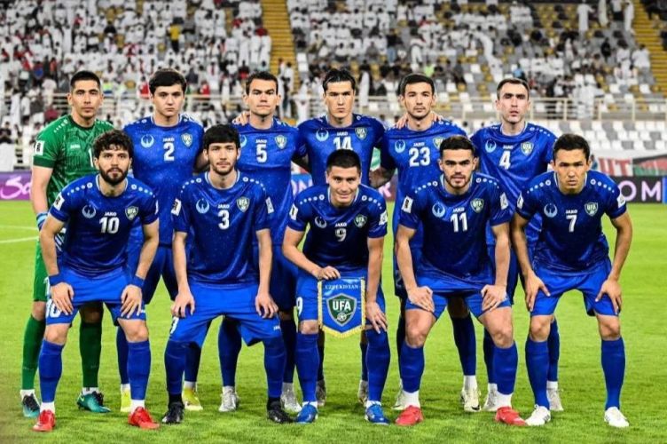 9fuzbek catrawarta Eleven soccer players in blue uniforms pose for a team photo on a green field with a goalkeeper in green on the far left and a crowd in the stands behind them