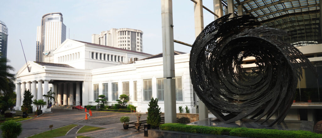 Bagiandepan2048x881 catrawarta White neoclassical museum with a columned portico a large black spiral sculpture in the foreground and a city skyline behind it
