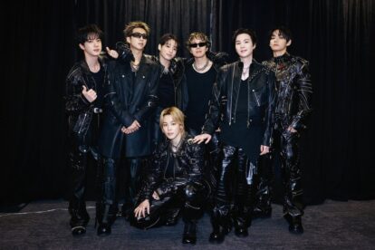 Seven men in black leather outfits posing together backstage against a dark curtain