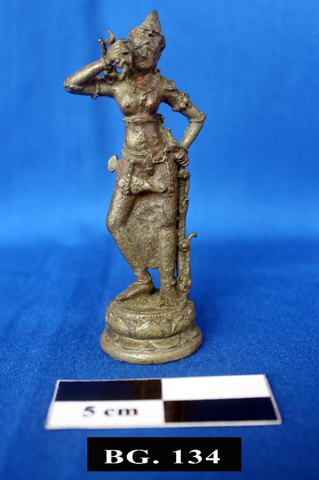 Bronze figurine of a standing female deity on a round base one hand on hip other near face next to a 5 cm scale against a blue backdrop