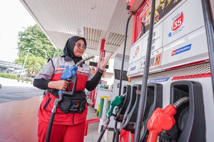 Harga bbm catrawarta Female gas station attendant in a red uniform and hijab holding a fuel nozzle while gesturing at a pump in a bright station setting