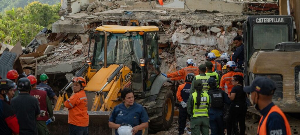 Rescue workers in orange vests gather around a yellow excavator amid a collapsed building and rubble at a disaster site in venezuela