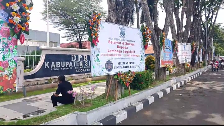 Row of banners along a tree lined sidewalk adorned with colorful flower arrangements outside a government complex A sign references kabupaten sidoarjo