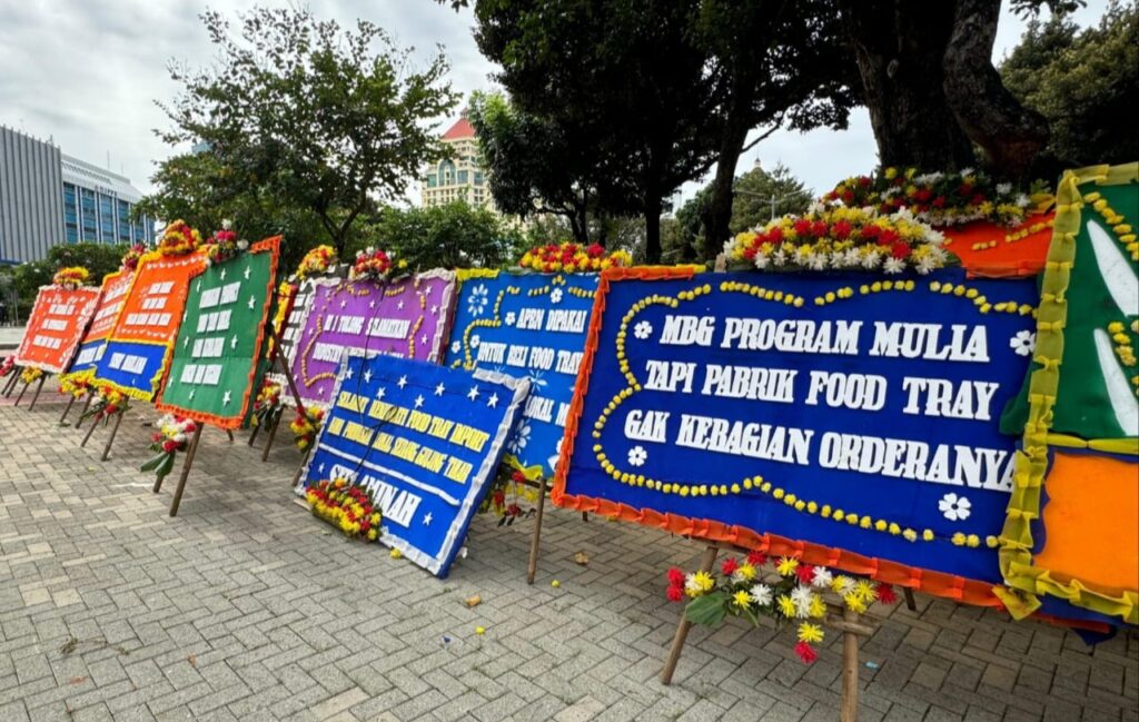 Row of large colorful condolence boards wrapped in flowers on stands along a paved walkway outside a parkbuilding
