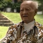 Old man with short white hair sitting in a garden wearing a brown and white tropical patterned shirt and a white turtleneck looking thoughtful
