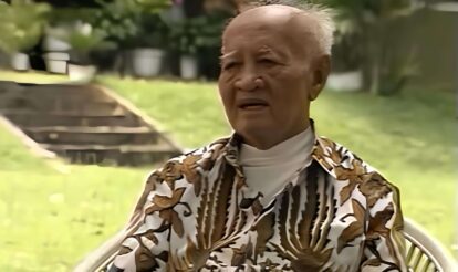 Old man with short white hair sitting in a garden, wearing a brown and white tropical-patterned shirt and a white turtleneck, looking thoughtful.