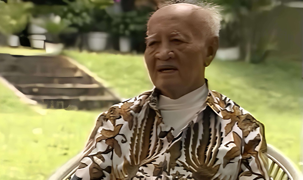 Old man with short white hair sitting in a garden wearing a brown and white tropical patterned shirt and a white turtleneck looking thoughtful