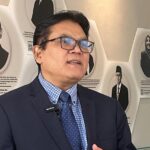 Man in a navy suit and glasses speaks wearing a lapel mic in front of a wall of black and white portrait plaques