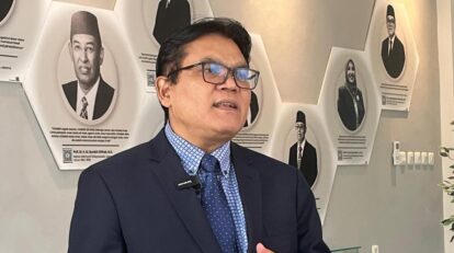 Man in a navy suit and glasses speaks, wearing a lapel mic, in front of a wall of black-and-white portrait plaques.