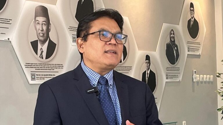 Man in a navy suit and glasses speaks, wearing a lapel mic, in front of a wall of black-and-white portrait plaques.