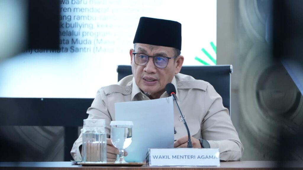 Indonesian official in a beige uniform and black cap speaks into a microphone at a conference with a'WAKIL MENTERI AGAMA' name sign on the desk.