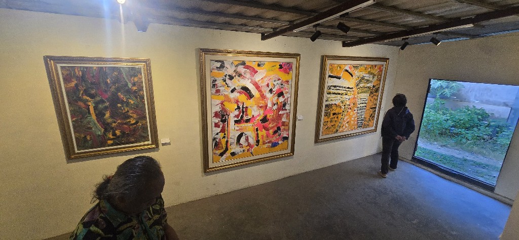 1002127341 catrawarta Two visitors view three framed abstract paintings in a gallery with a window showing greenery on the right side