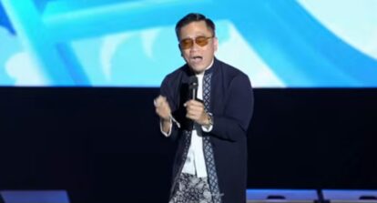 Male performer on a stage, speaking into a microphone, wearing sunglasses, a dark coat and patterned scarf, with a blue abstract backdrop behind him.