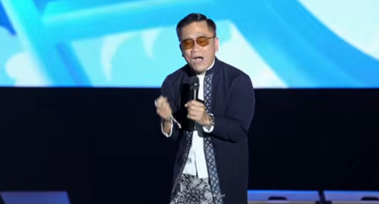 Gus miftah catrawarta Male performer on a stage speaking into a microphone wearing sunglasses a dark coat and patterned scarf with a blue abstract backdrop behind him