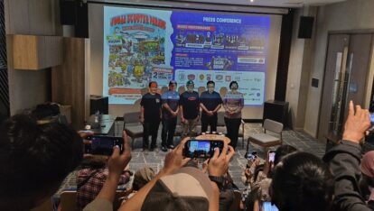 Group of five people standing on a stage in front of a large poster during a press conference, audience taking photos in a conference room.