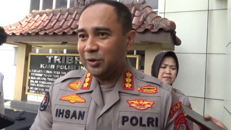 Polri police officer named Ihsan speaking to reporters outdoors with a signboard behind him.