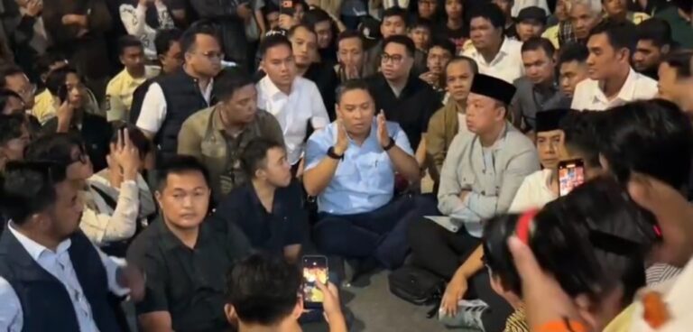Man in a light blue shirt gesturing while speaking to a large seated crowd; many people recording with phones.