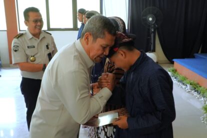 Two men bow and clasp forearms in a formal handshake during an awards ceremony a uniformed officer watches smiling in the background indoors