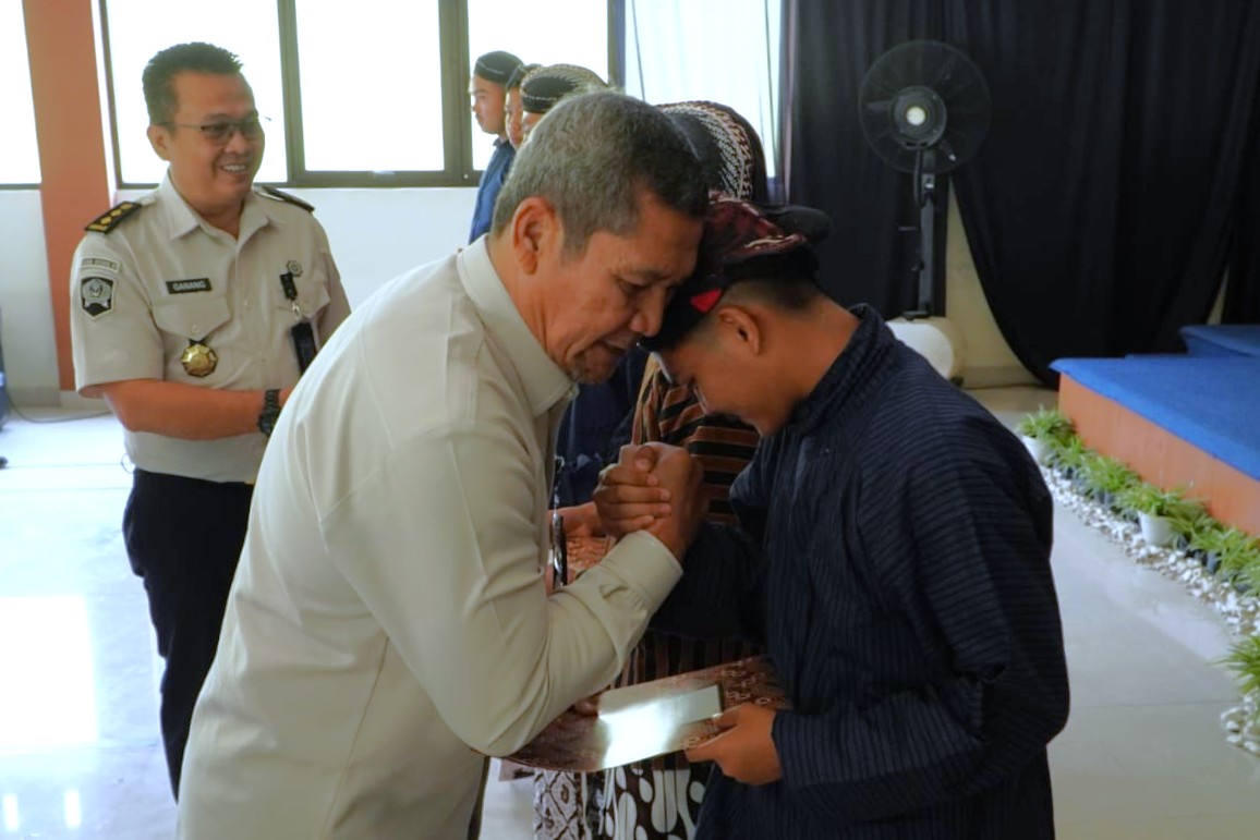 Penyerahan surat putusan pengurangan masa pidana bagi anak catrawarta Two men bow and clasp forearms in a formal handshake during an awards ceremony a uniformed officer watches smiling in the background indoors