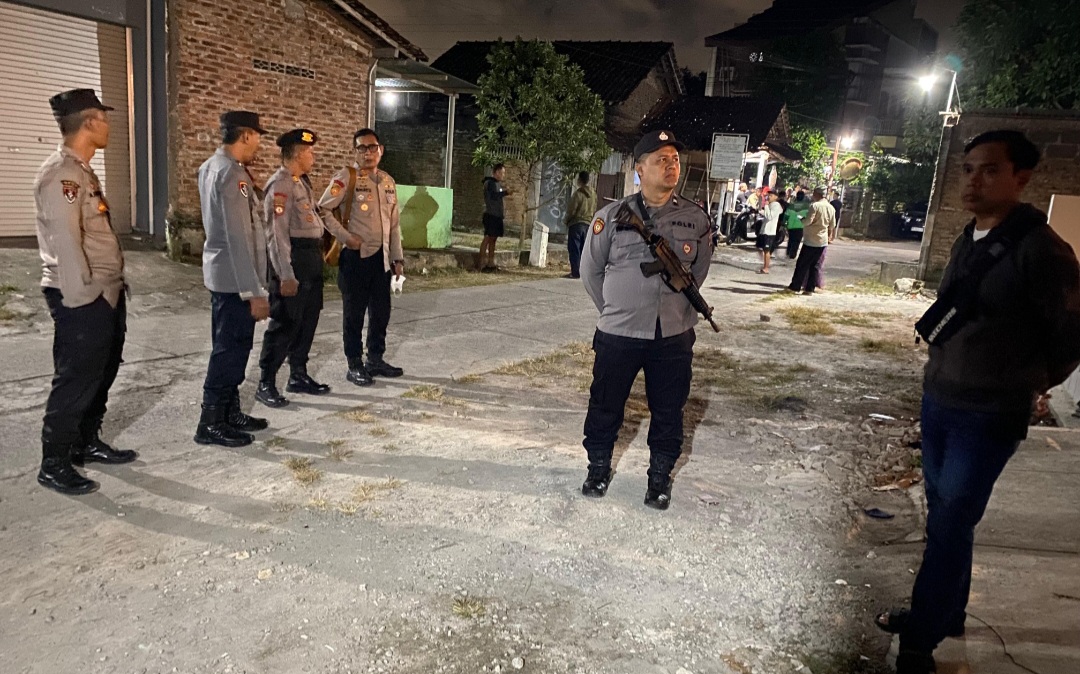Indonesian police officers stand in a line on a dusty street at night one holding a rifle with bystanders in the background beside brick buildings