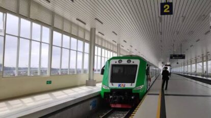 Front view of a green commuter train stopped at platform 2 in a bright modern station with large windows a person stands on the platform nearby contrast