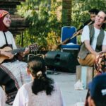 Musician in a colorful headscarf plays a small guitar while an older man with a headset sings on an outdoor stage surrounded by a small audience