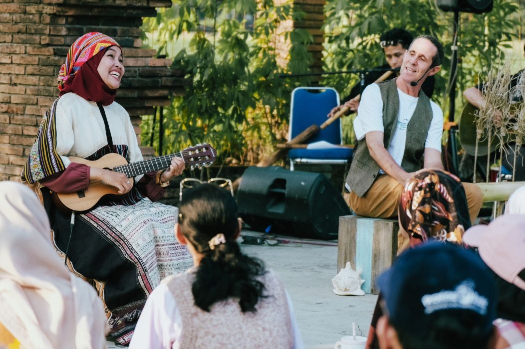 10gdongeng1 catrawarta Musician in a colorful headscarf plays a small guitar while an older man with a headset sings on an outdoor stage surrounded by a small audience
