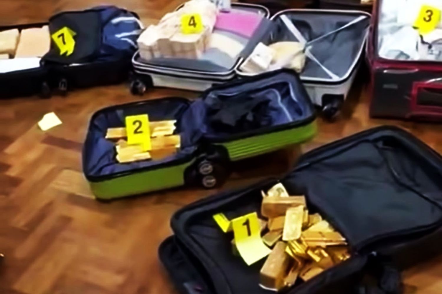 Open suitcase on a wooden floor filled with gold bars stacked inside labeled with yellow evidence tags 1 2 3 4 Other suitcases surround it