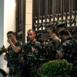 Group of soldiers in camouflage uniforms with rifles standing near a decorative wrought iron fence outside a building