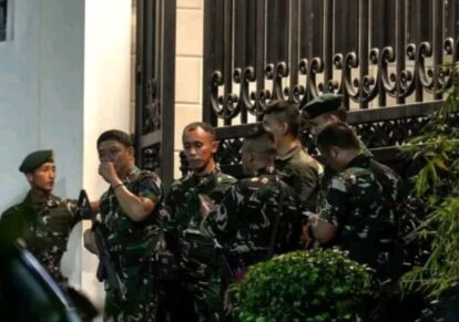Group of soldiers in camouflage uniforms with rifles, standing near a decorative wrought-iron fence outside a building.