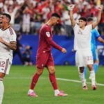 Soccer players celebrate a goal on the field with arms raised and fans cheering in the stands behind them