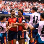 Group of spain players celebrate a goal on the pitch wearing red jerseys and pink world cup 2026 bibs in a crowded stadium
