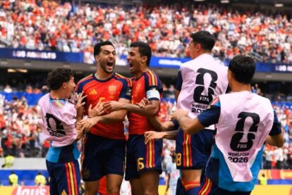 Group of Spain players celebrate a goal on the pitch, wearing red jerseys and pink World Cup 2026 bibs, in a crowded stadium.