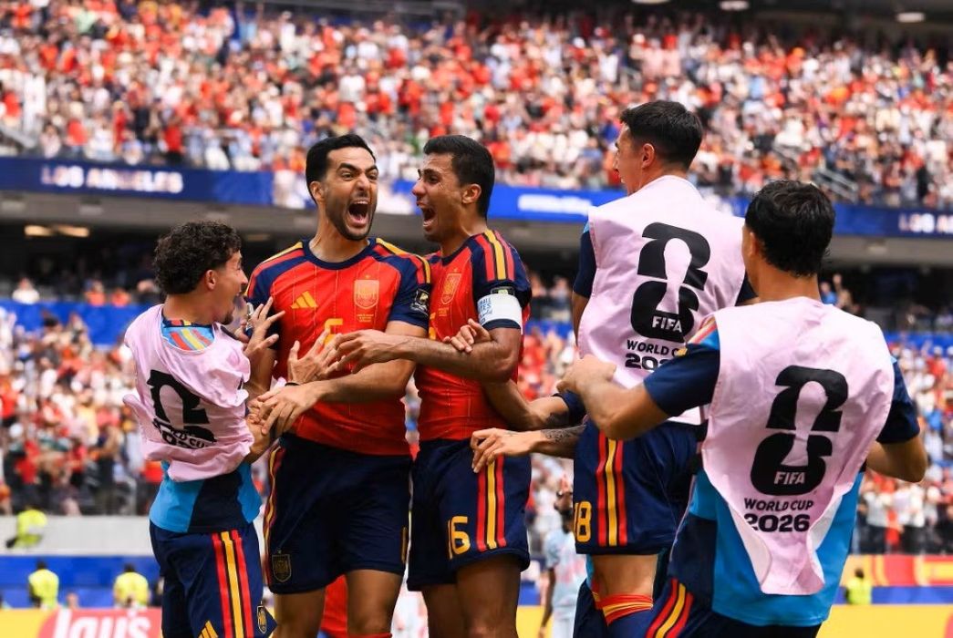 11gspain1 catrawarta Group of spain players celebrate a goal on the pitch wearing red jerseys and pink world cup 2026 bibs in a crowded stadium