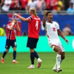England forward no 10 celebrates a goal with a raised fist as teammates in red react in the background on a soccer field in front of cheering fans